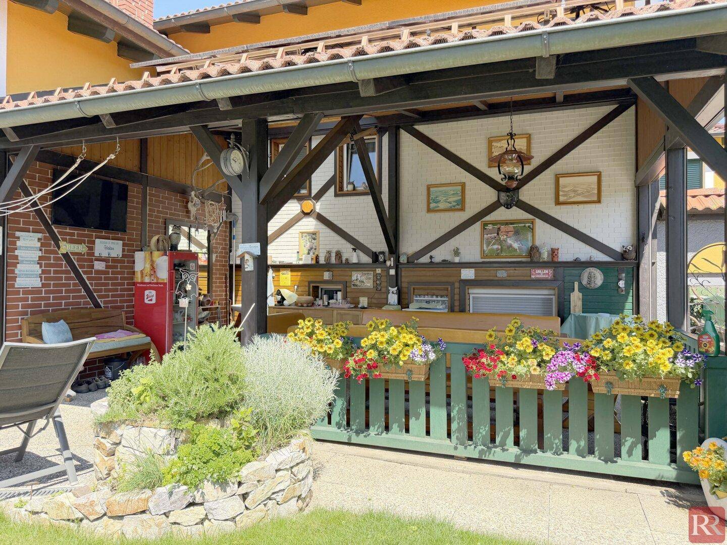 Überdachte Terrasse mit Holzbalken, Blumenkästen und gemütlicher Sitzecke im Freien.
