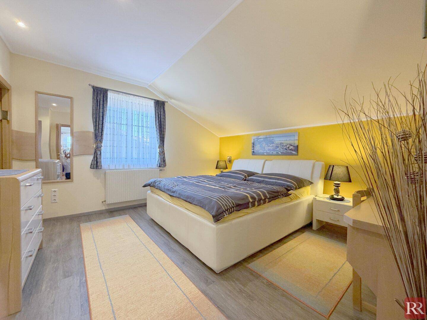Schlafzimmer mit gelber Akzentwand, Doppelbett und modernem Laminatboden.