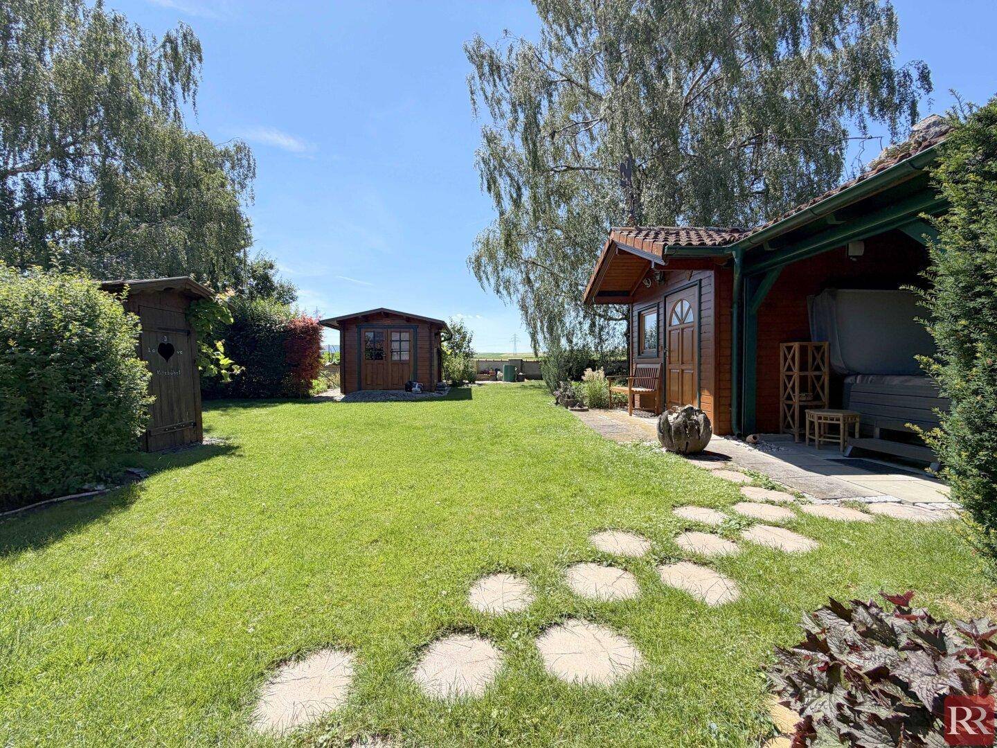 Gepflegter Garten mit Rasen, Trittsteinen und kleinen Gartenhäusern im Hintergrund.