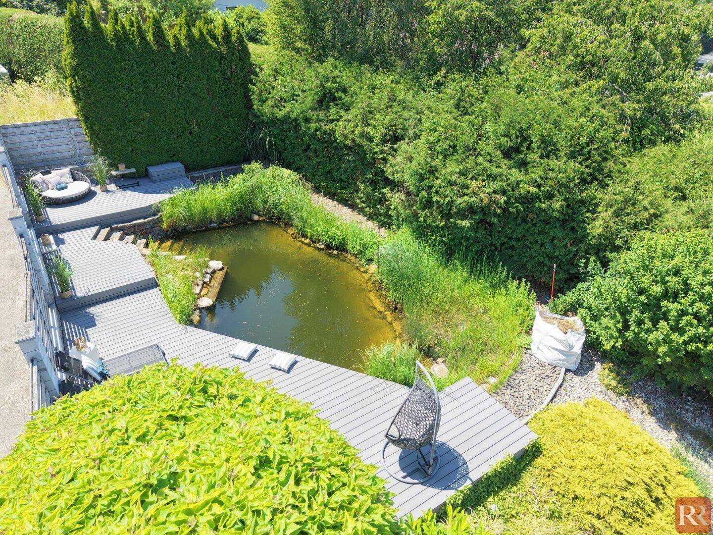 Natürlicher Gartenteich mit Holzdeck und Sitzgelegenheiten, umgeben von üppigem Grün.