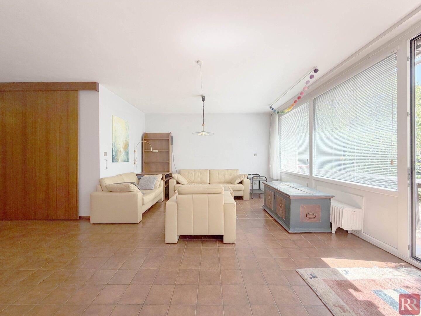 Helles Wohnzimmer mit großen Fenstern, Fliesenboden und beigen Sofas.