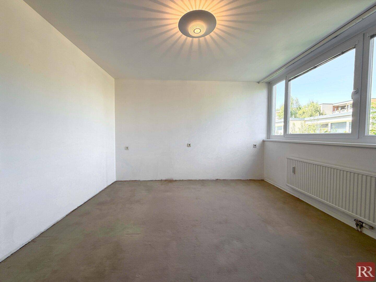 Leerer Raum mit großem Fenster, Heizkörper und unfertigem Bodenbelag.