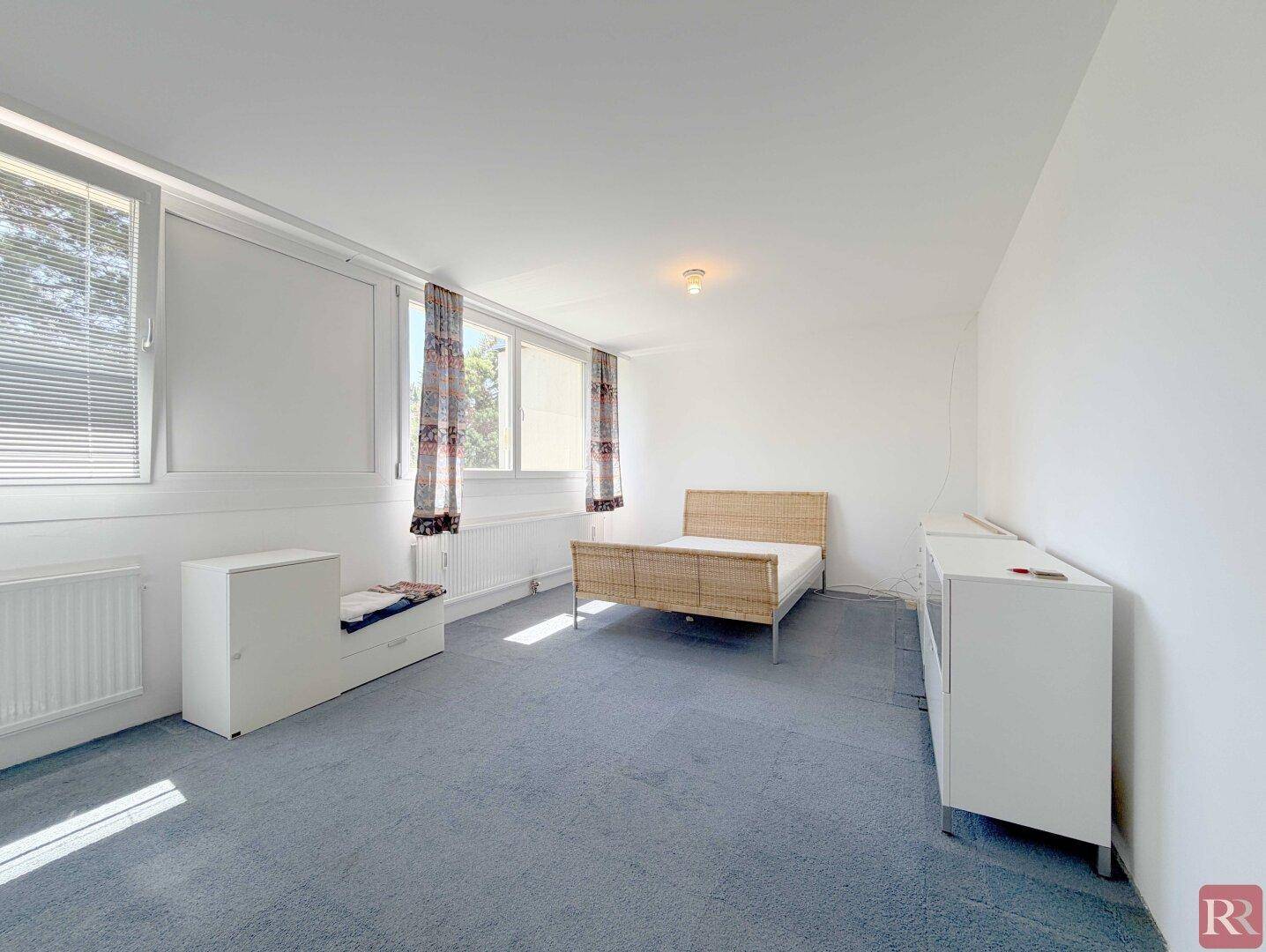 Schlafzimmer mit blauem Teppichboden, Doppelbett und großen Fenstern mit Jalousien.
