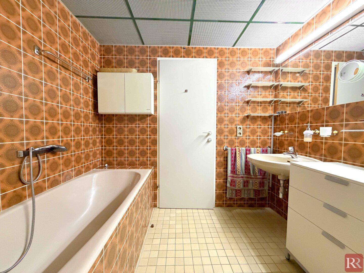 Badezimmer im 80er-Jahre-Stil mit braunen Fliesen, Badewanne und Einzelwaschbecken.