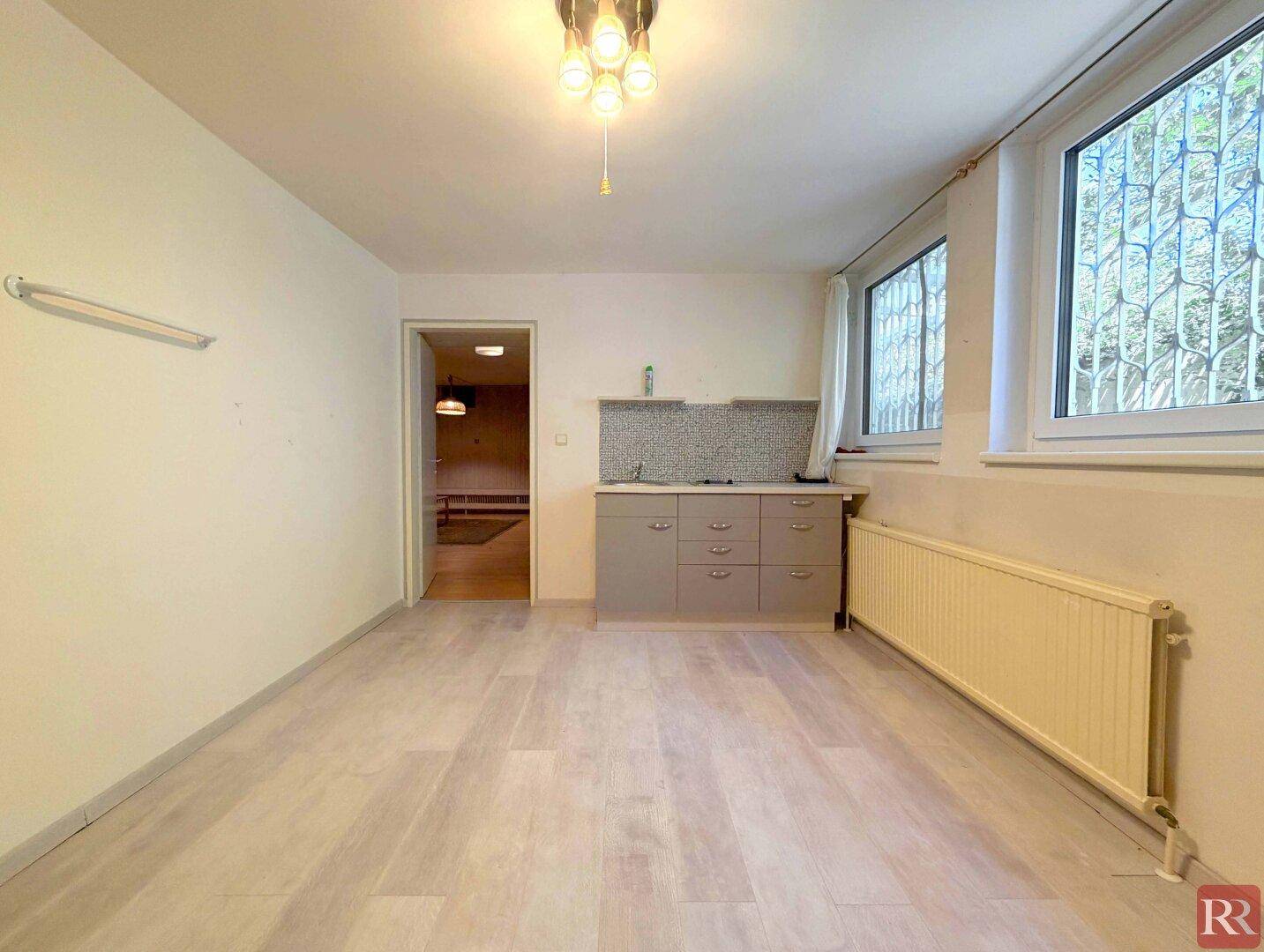 Kellerraum mit heller Holzoptik-Boden, kleiner Küchenzeile und vergitterten Fenstern.