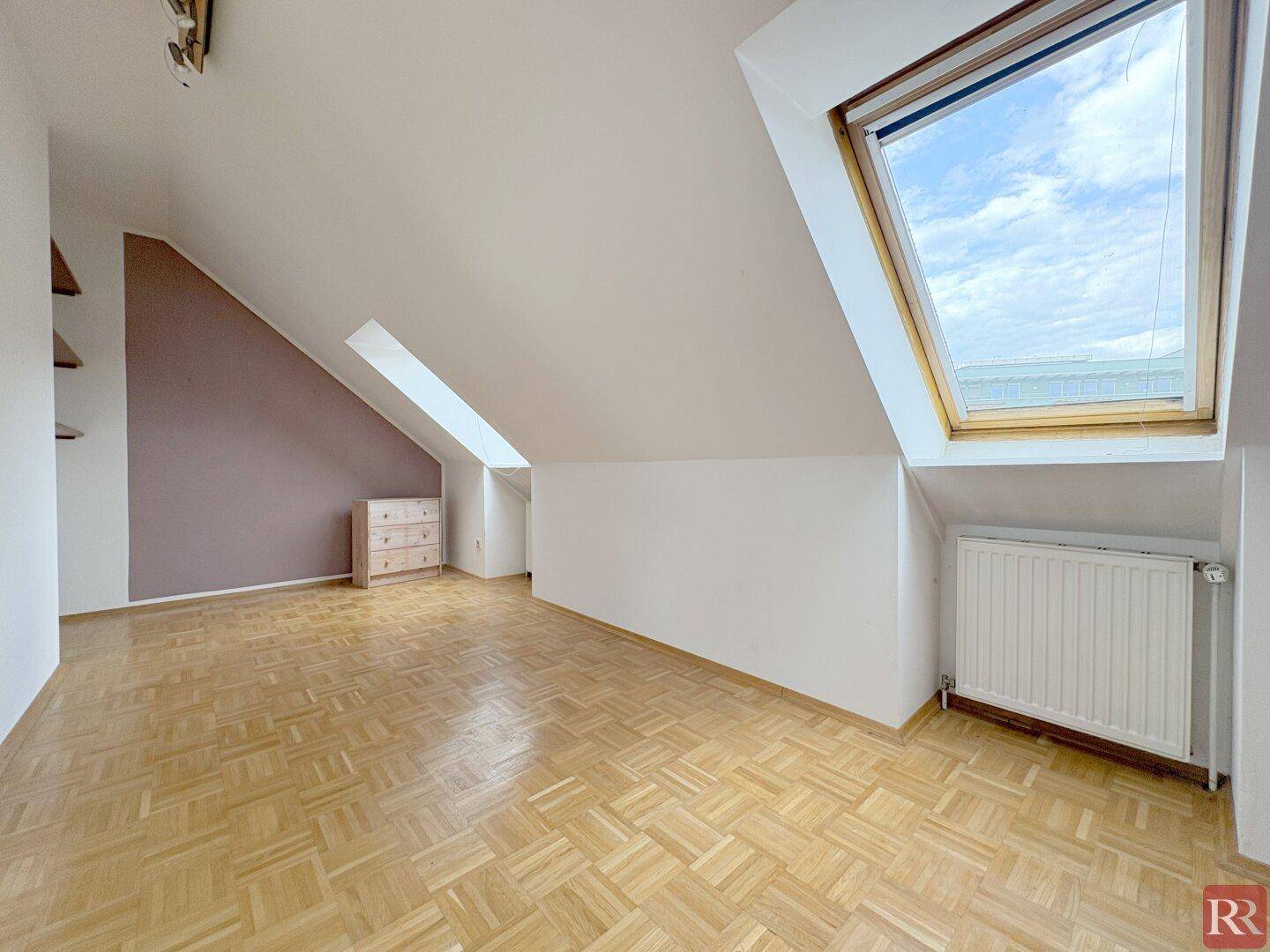 Geräumiges Zimmer mit Parkettboden, Dachschrägen und einem großen Dachfenster, das den Raum hell erleuchtet.