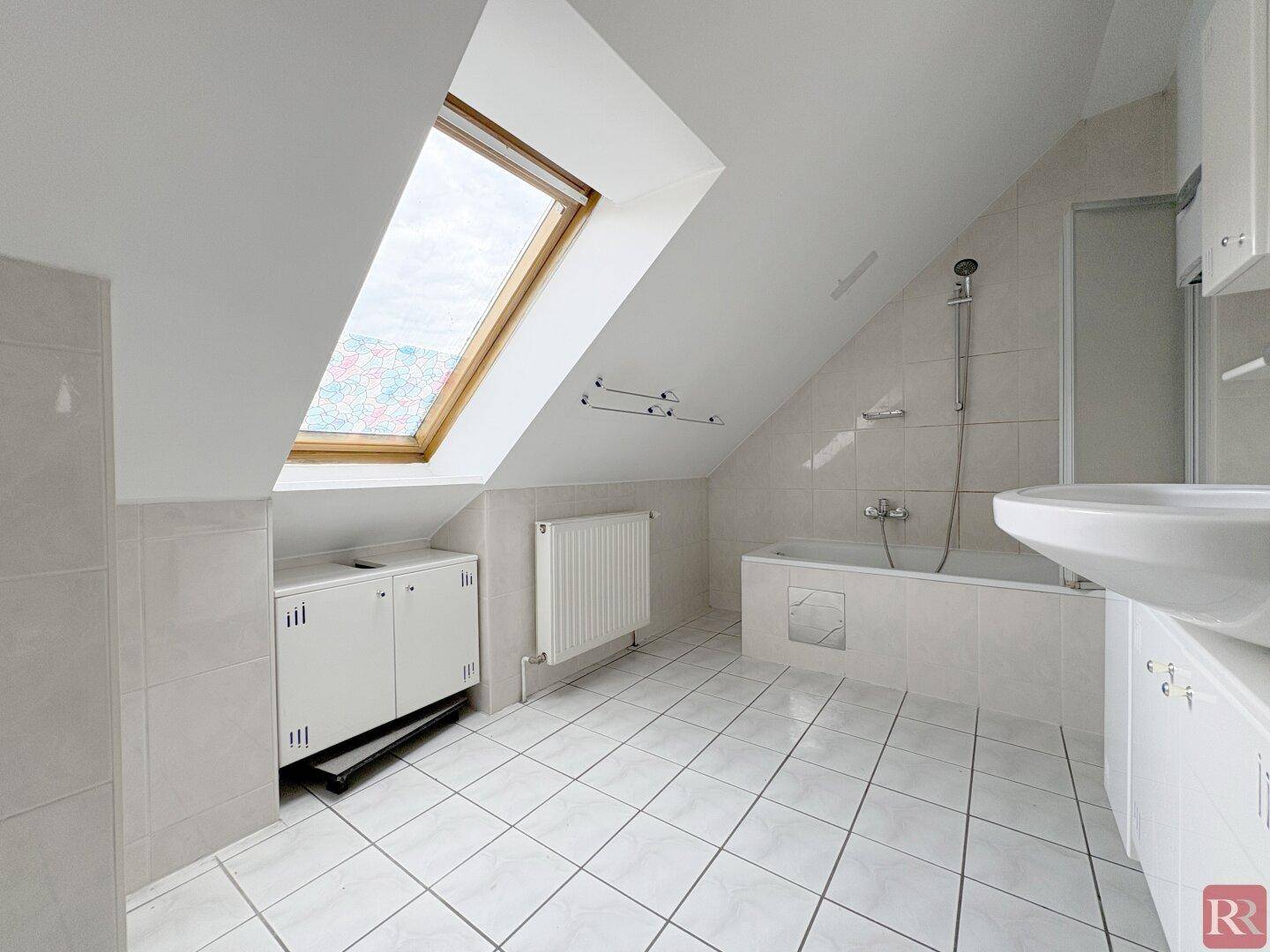 Helles Badezimmer mit Badewanne, Waschbecken, weißen Fliesen und einem großen Dachfenster in der Schräge.