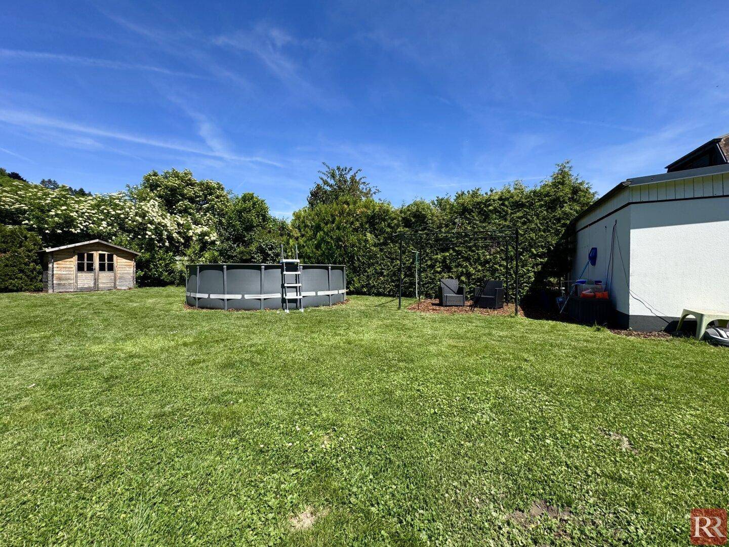 Sonniger Garten mit Aufstellpool, kleiner Sitzecke und Gartenhaus, umgeben von hohen Hecken.