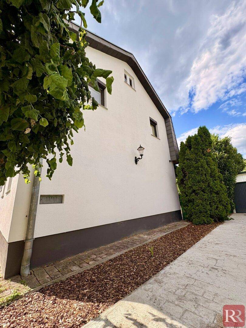 Seitenansicht des Hauses mit weißer Fassade, kleinen Fenstern und gepflastertem Weg.