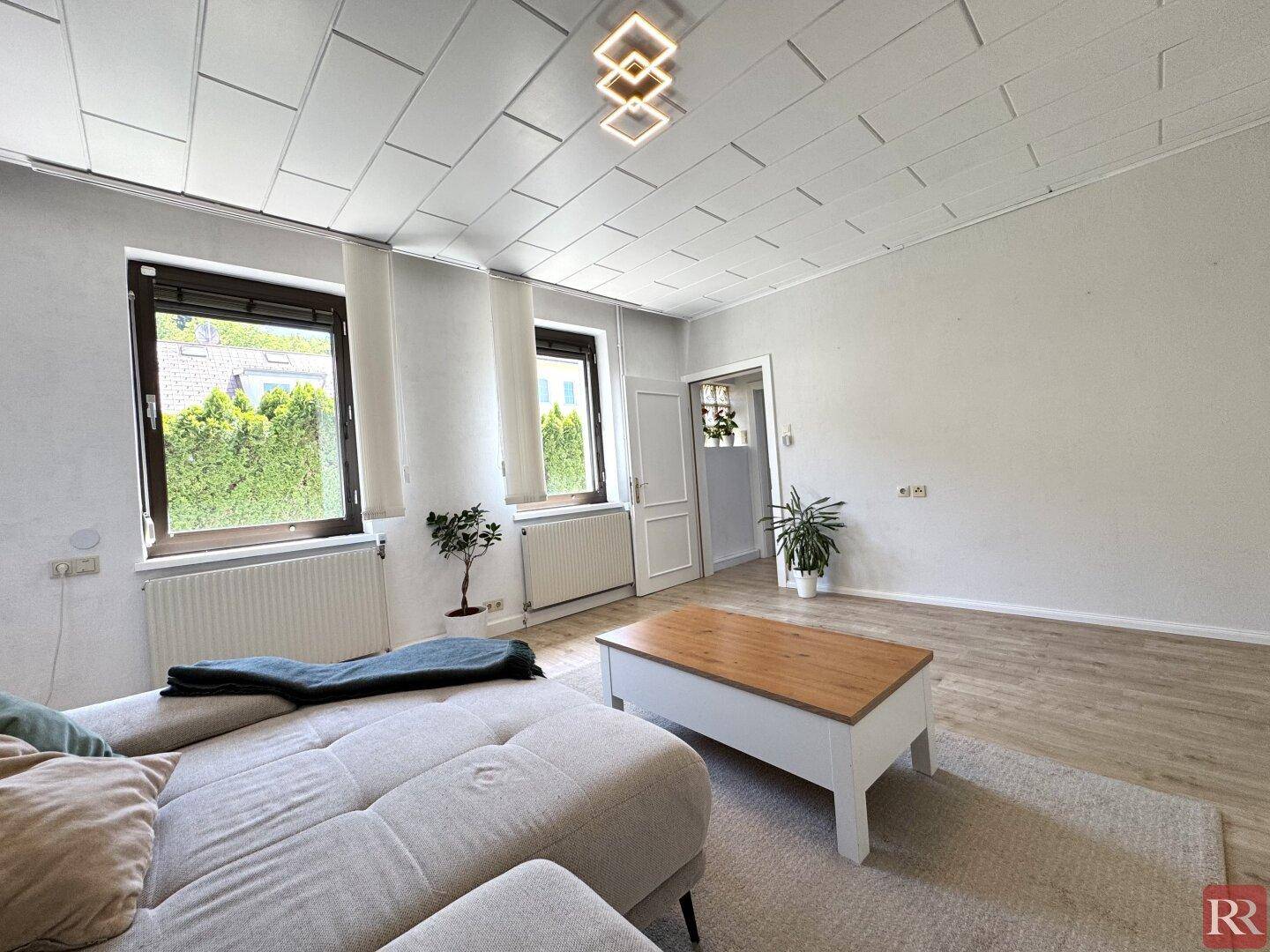 Freundliches Wohnzimmer mit zwei Fenstern, Heizkörpern und Zugang zu einem weiteren Raum.