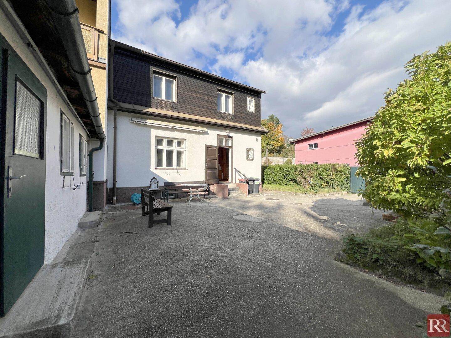 Gepflasterter Hofbereich neben dem Haus mit Sitzbank und Nebengebäude an einem sonnigen Tag.