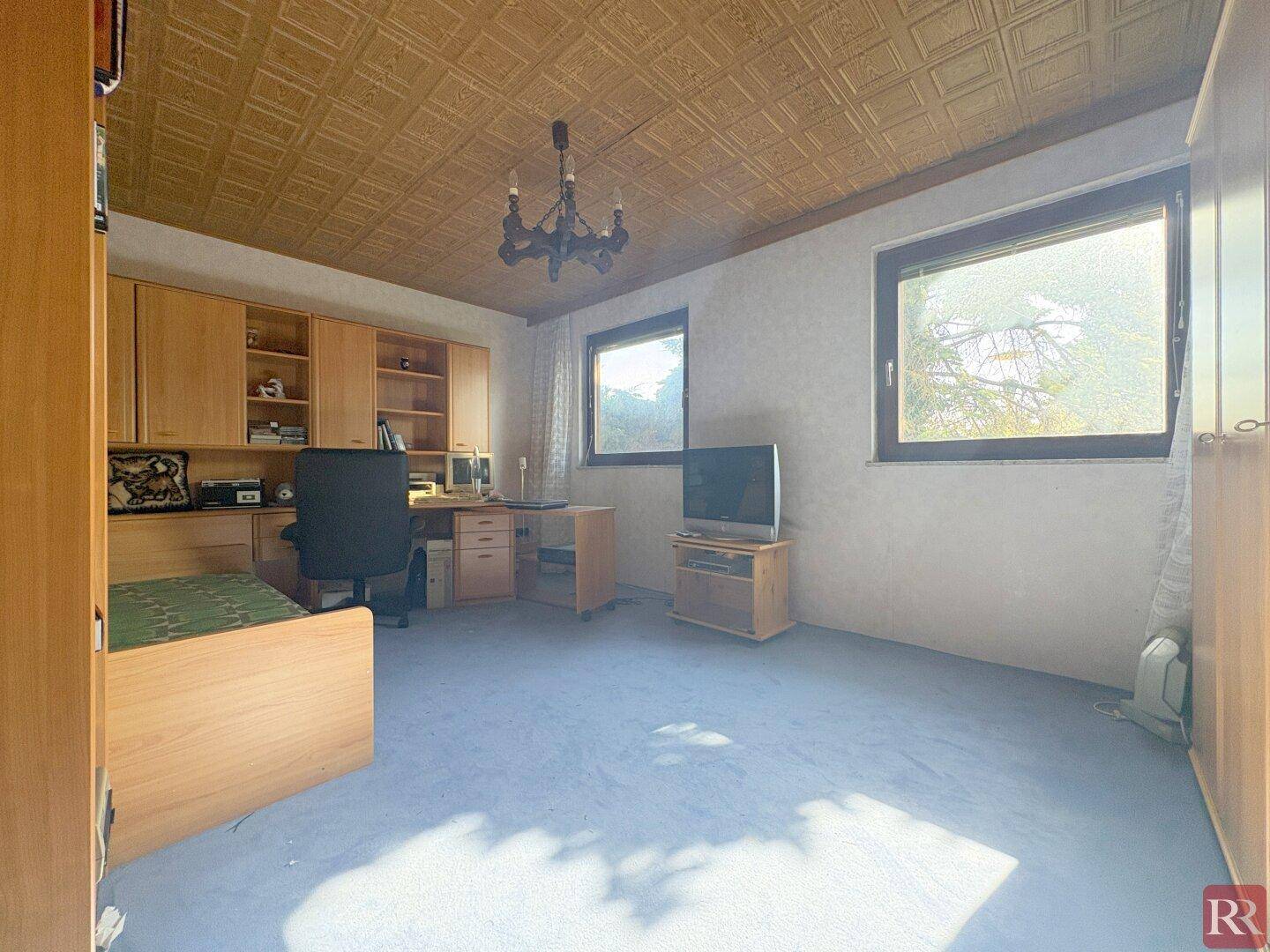 Helles Arbeitszimmer mit blauem Teppichboden, Holzmöbeln und zwei Fenstern.