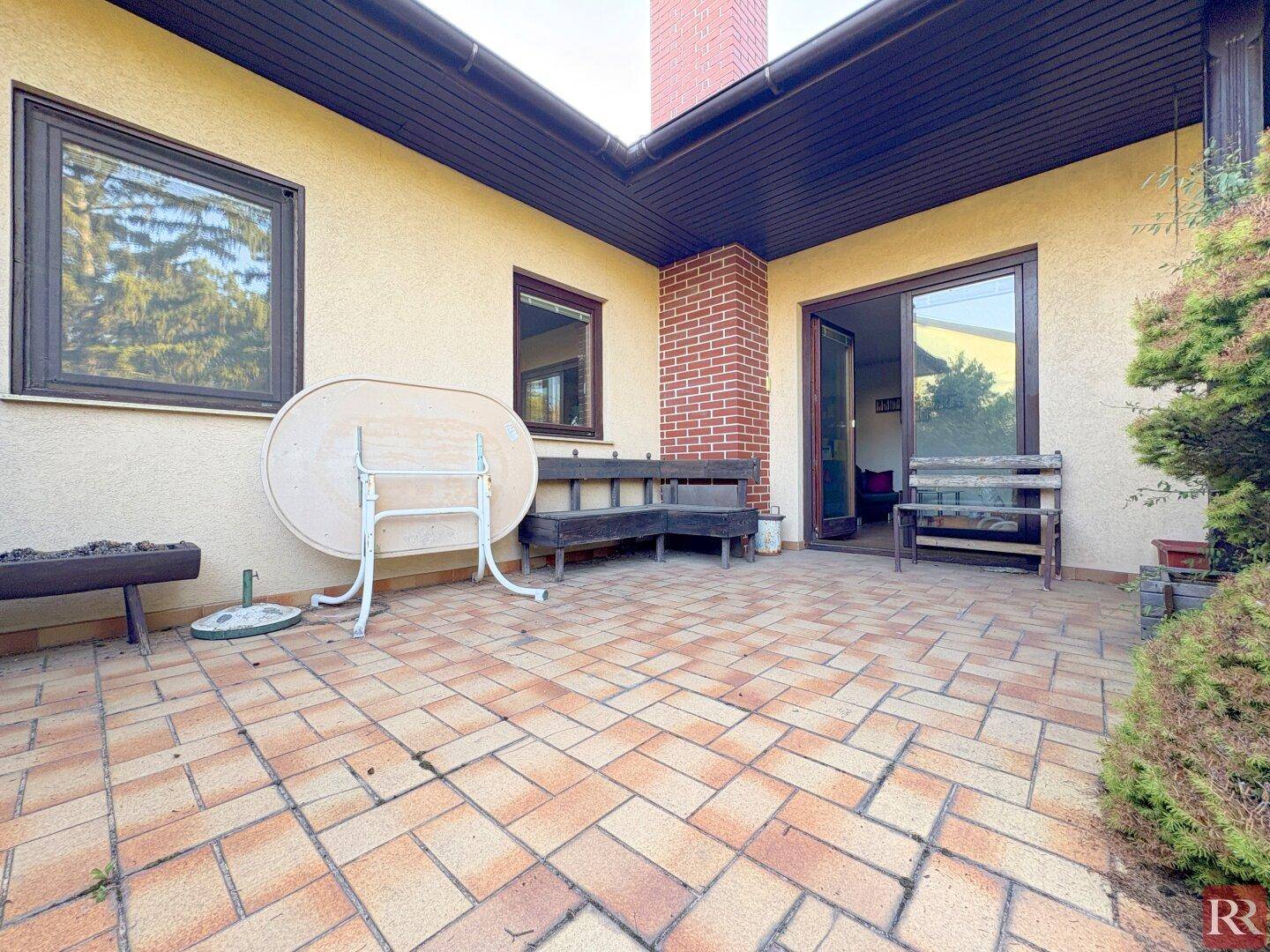 Gedeckte Terrasse mit Ziegelboden, Sitzbänken und Zugang zum Garten.