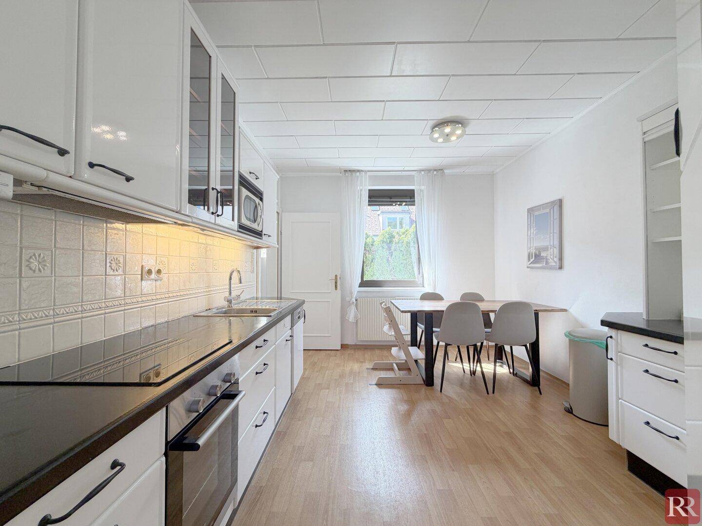 Moderne Einbauküche mit weißen Schränken, dunkler Arbeitsplatte und Essbereich am Fenster.