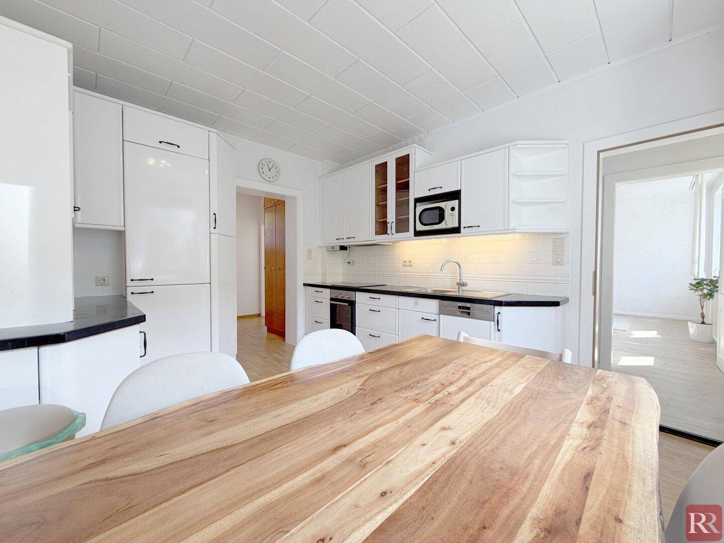 Geräumige Küche mit hellem Holztisch, weißen Schränken und Blick in den angrenzenden Wohnbereich.