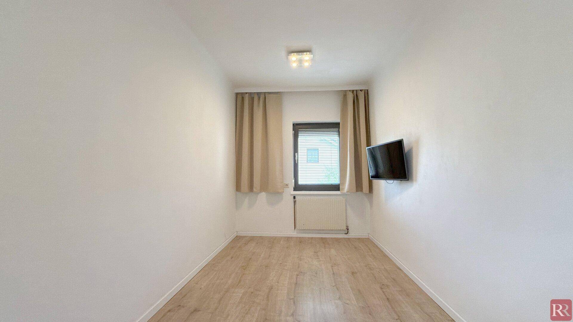 Schlafzimmer mit Laminatboden, einem Fenster und einem an der Wand montierten Fernseher.