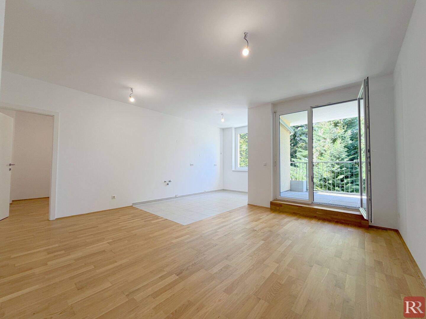 Heller Wohnbereich mit Parkettboden und Zugang zu einem Balkon, umgeben von Bäumen.