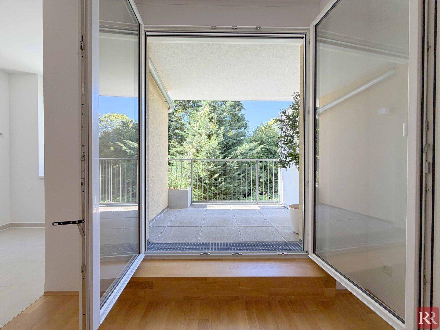 Blick vom Wohnzimmer auf den gefliesten Balkon mit Metallgeländer und grüner Umgebung.