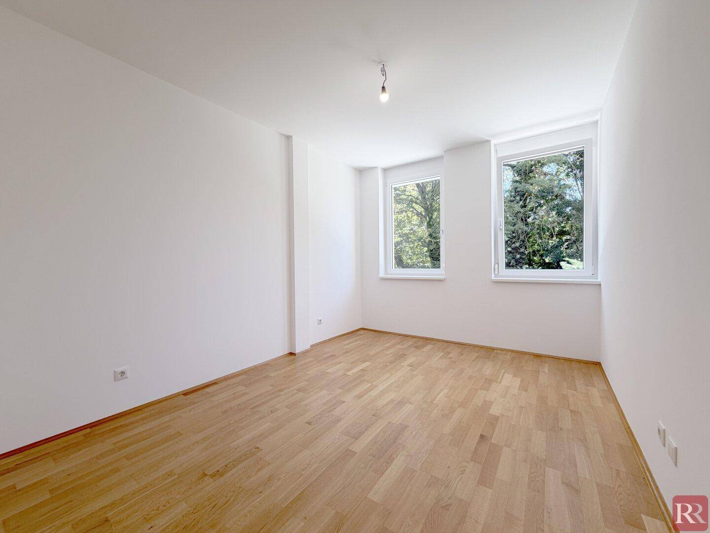 Helles Schlafzimmer mit Parkettboden und zwei Fenstern mit Blick ins Grüne.