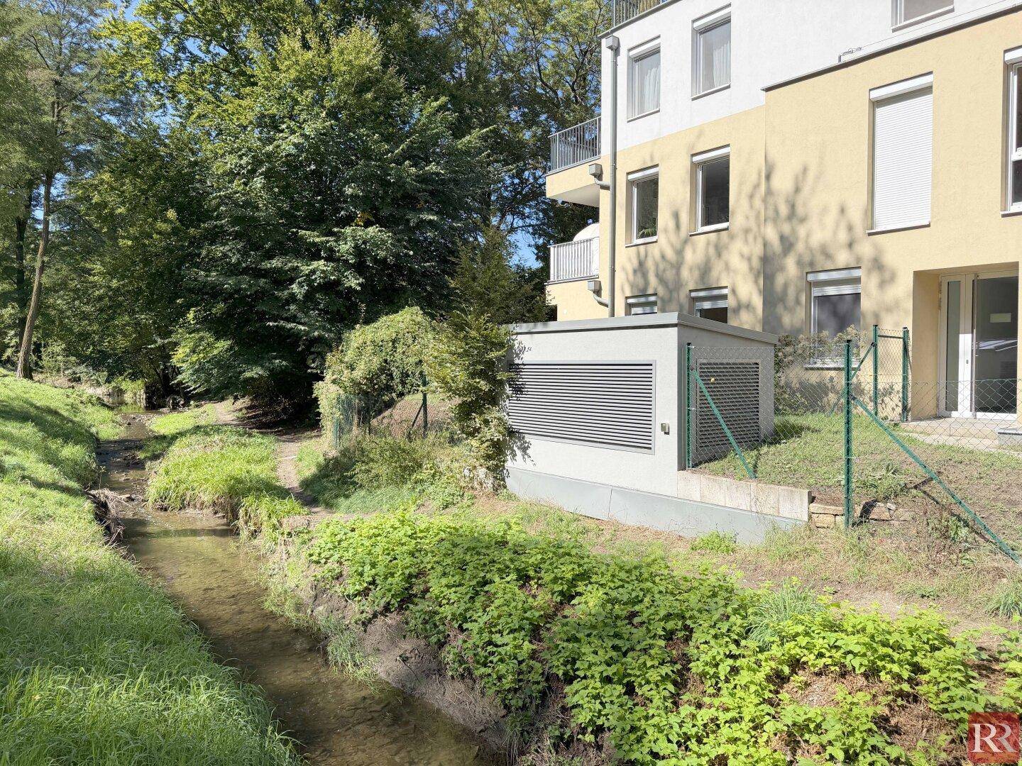Ansicht des modernen Gebäudes von der Bachseite mit viel Grün und sonniger Lage.