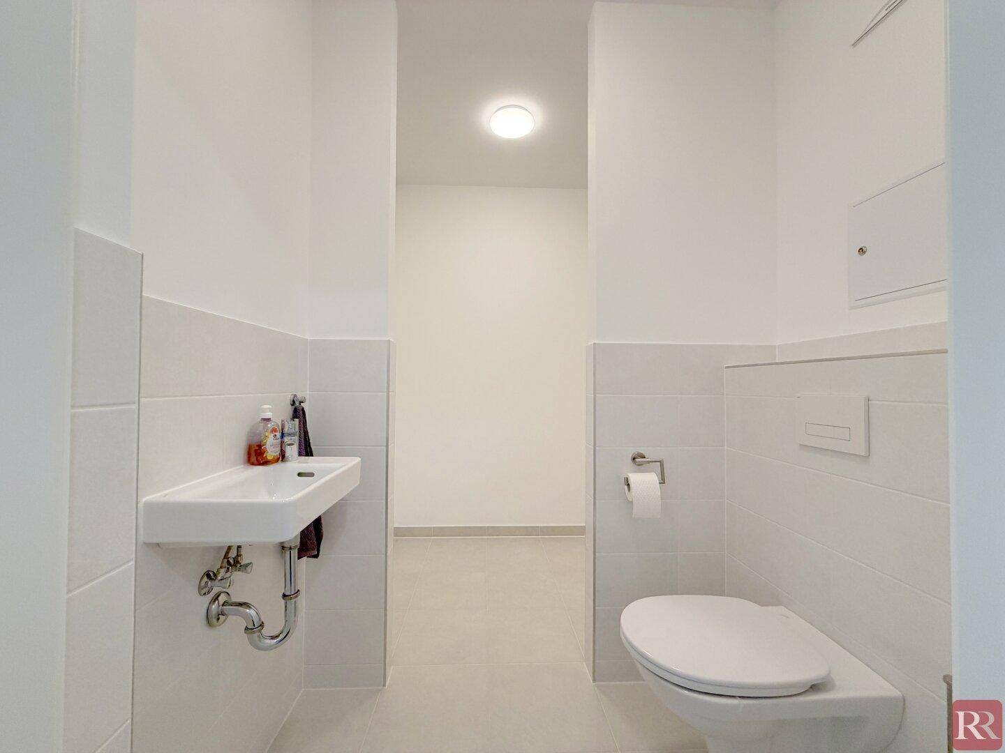 Funktionale Gästetoilette mit weißen Fliesen, WC und kleinem Handwaschbecken.