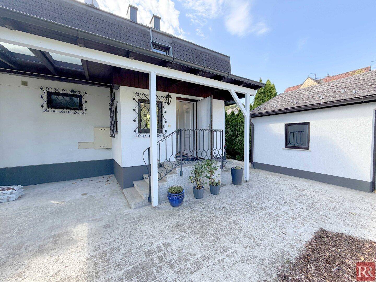 Überdachter Eingangsbereich mit gepflastertem Boden und dekorativen Pflanzen, der zum Haus führt.