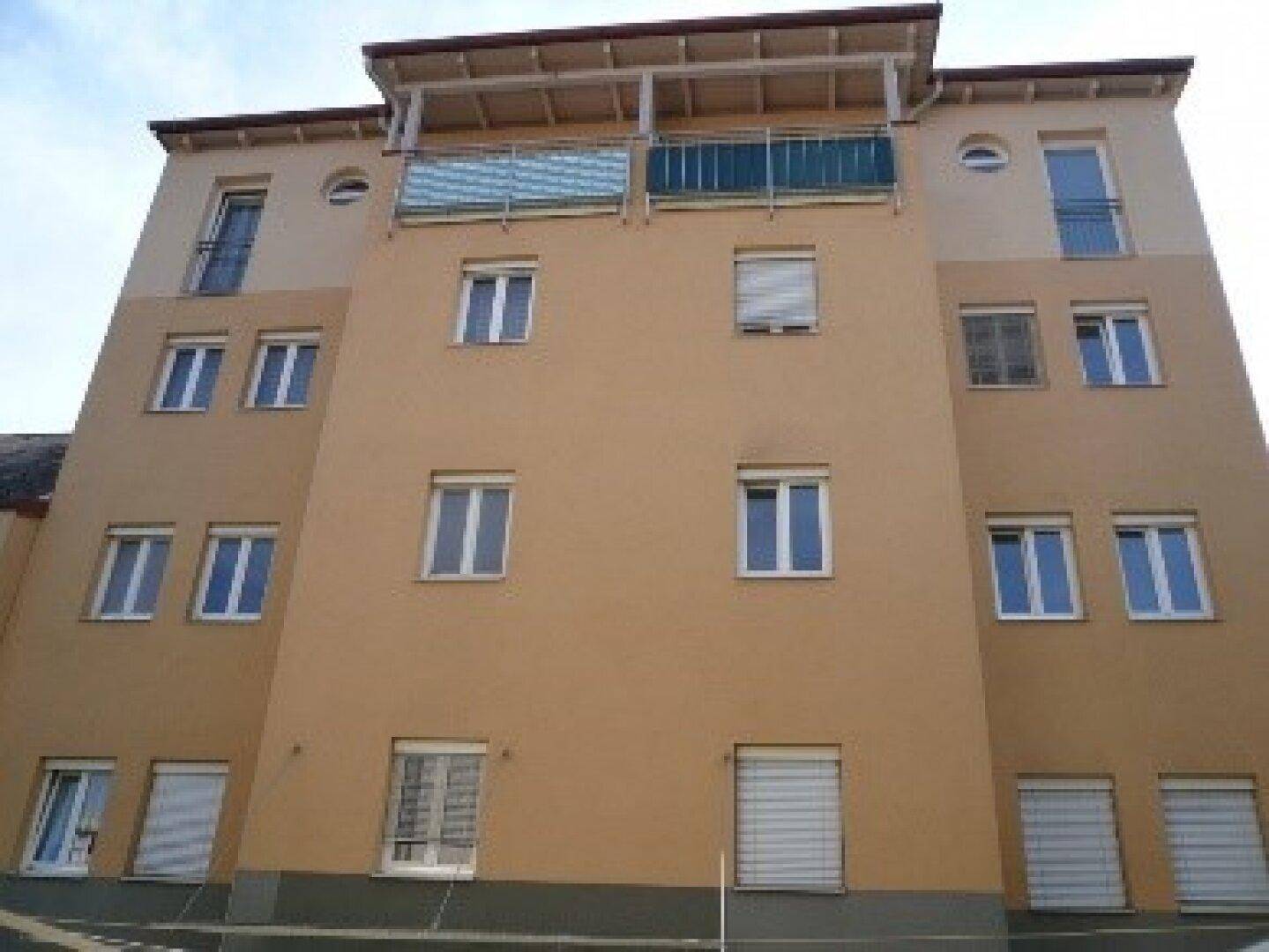 Mehrstöckiges Wohngebäude mit beiger Fassade, weißen Fenstern und Balkonen im oberen Bereich.