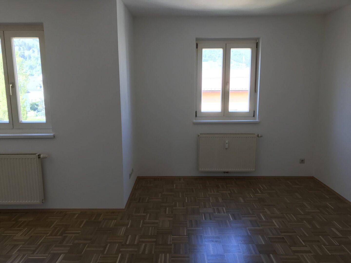 Geräumiger, heller Raum mit Parkettboden, weißen Wänden und zwei großen Fenstern.
