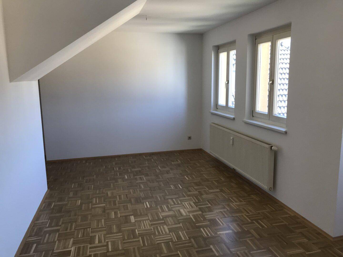 Heller Raum mit Parkettboden, Dachschrägen und zwei Fenstern, die viel Tageslicht hereinlassen.