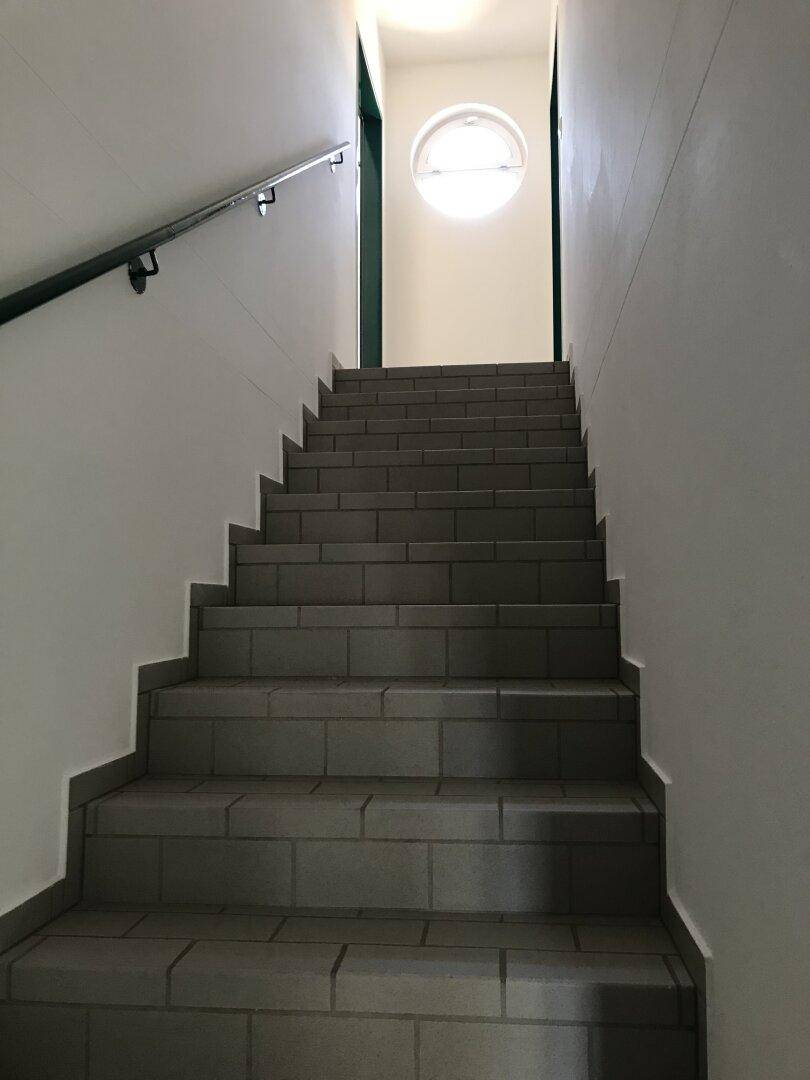 Geflieste Treppe mit Handlauf und einem runden Fenster am oberen Ende, das Licht spendet.