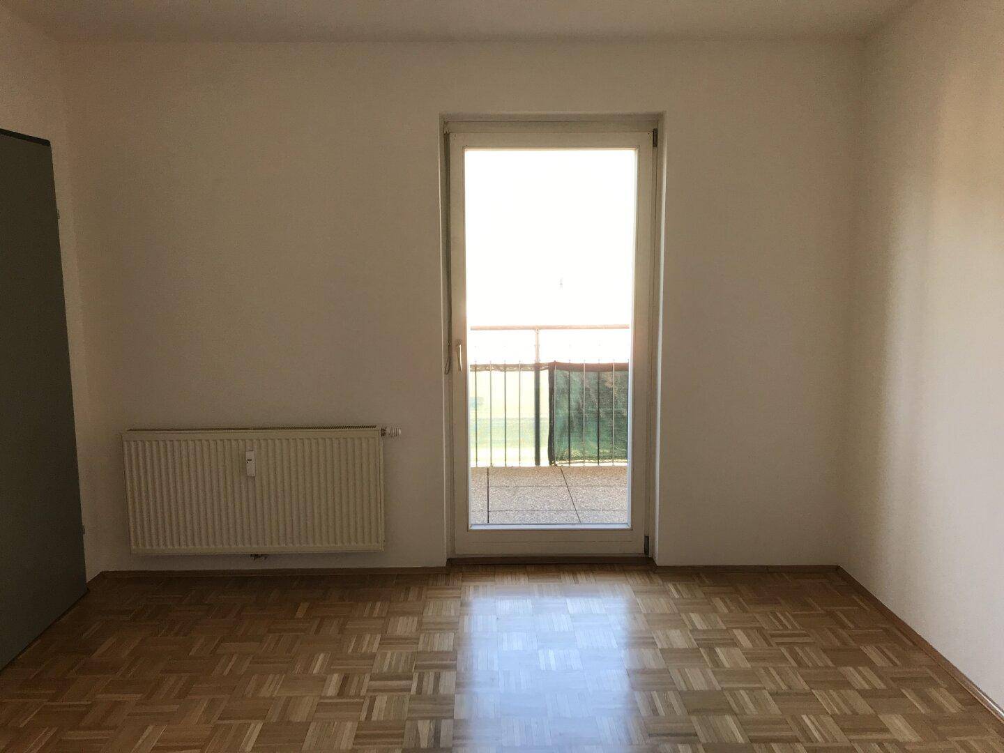 Heller, leerer Raum mit Parkettboden, weiß gestrichenen Wänden und Zugang zu einem Balkon.