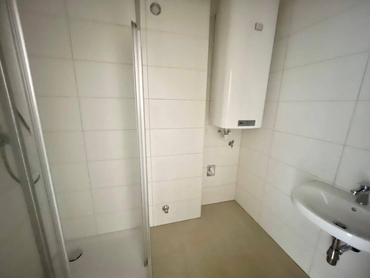 Funktionales Badezimmer mit weißen Fliesen, Duschkabine, Waschbecken und Warmwasserboiler.