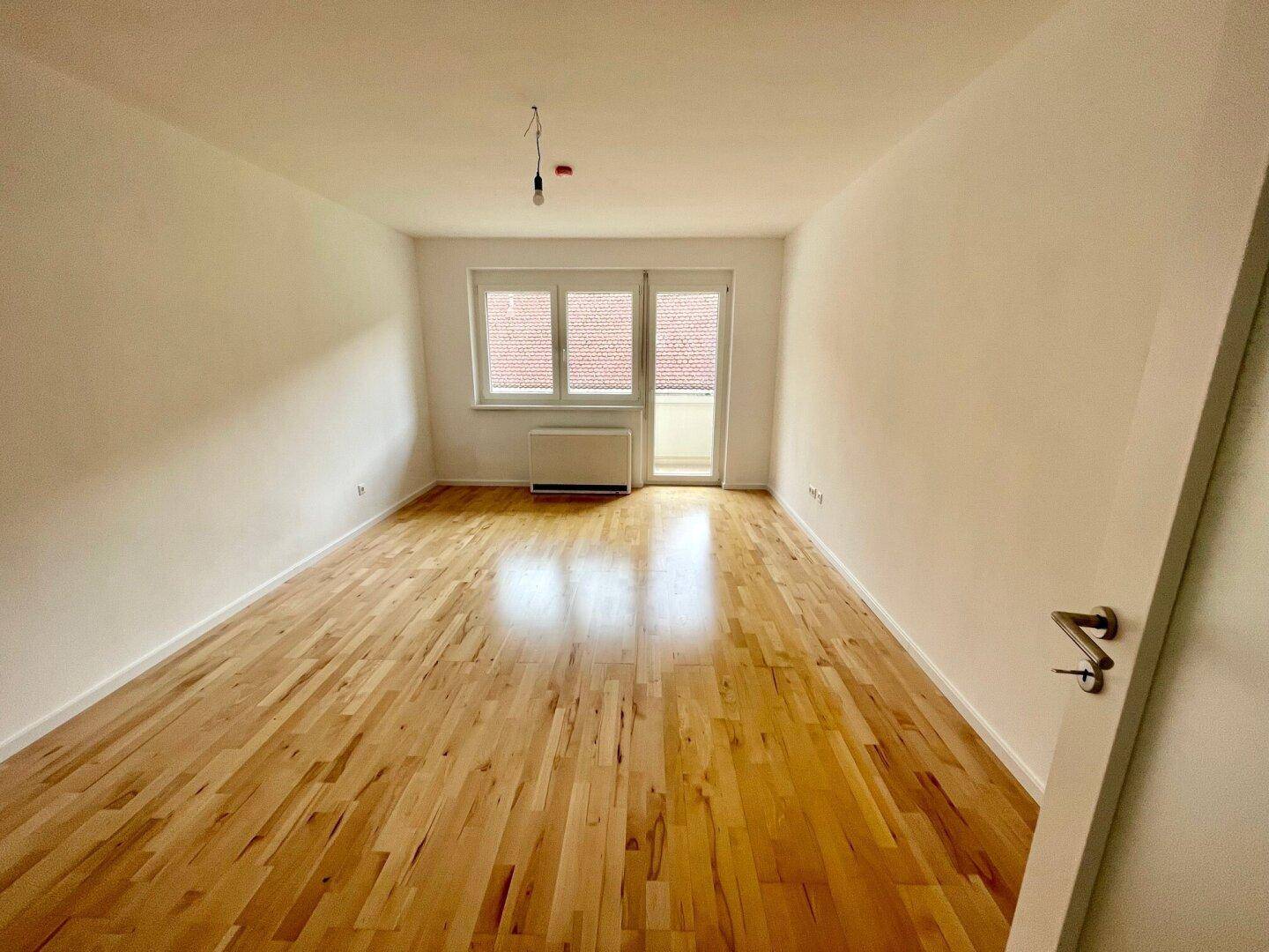 Geräumiger, heller Raum mit Holzboden, großen Fenstern und Zugang zu einem kleinen Balkon.