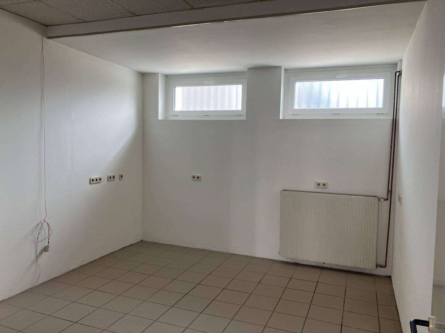 Kleinerer Raum mit zwei Fenstern, Fliesenboden und einem Heizkörper, ideal als Büro oder Lager.