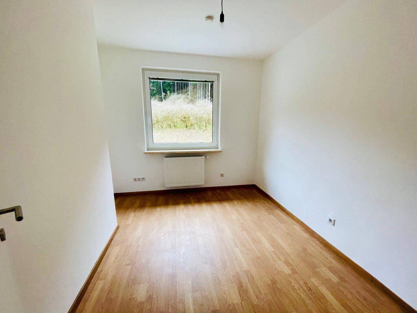 Helles Schlafzimmer mit Holzparkettboden, weißen Wänden und einem Fenster mit Blick auf die Natur.