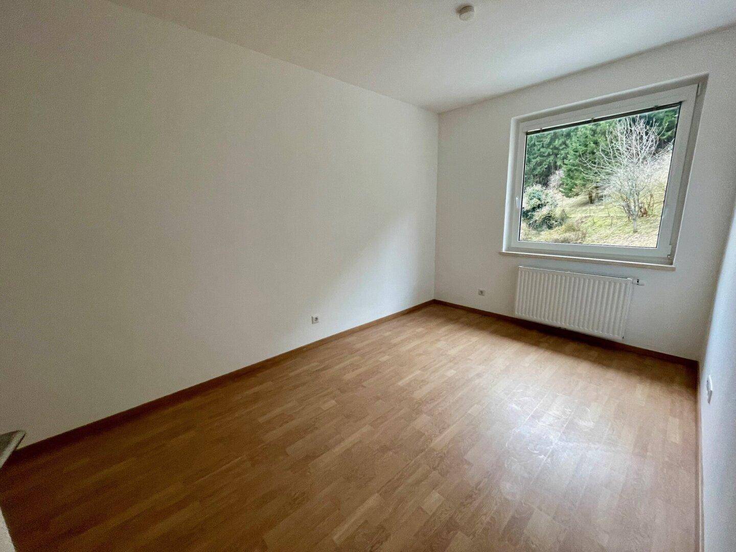 Geräumiges Zimmer mit Holzboden, weißen Wänden und einem großen Fenster mit Blick auf die bewaldete Umgebung.