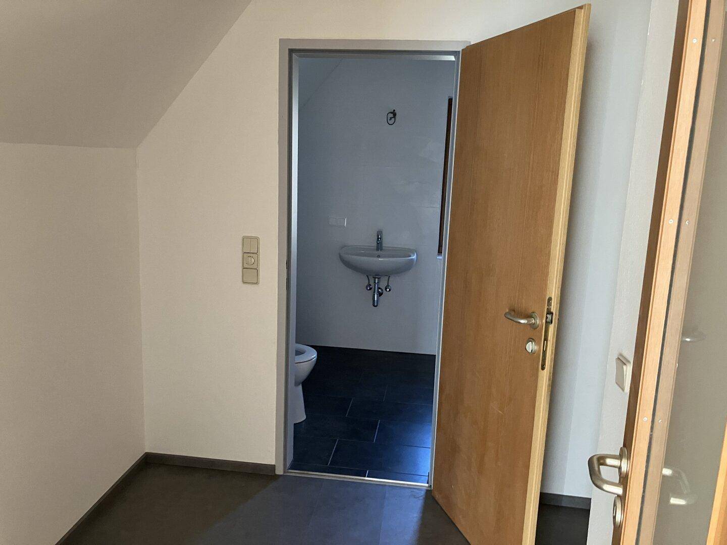 Flur mit dunklem Fliesenboden und offener Tür zu einem Badezimmer mit WC und Waschbecken.