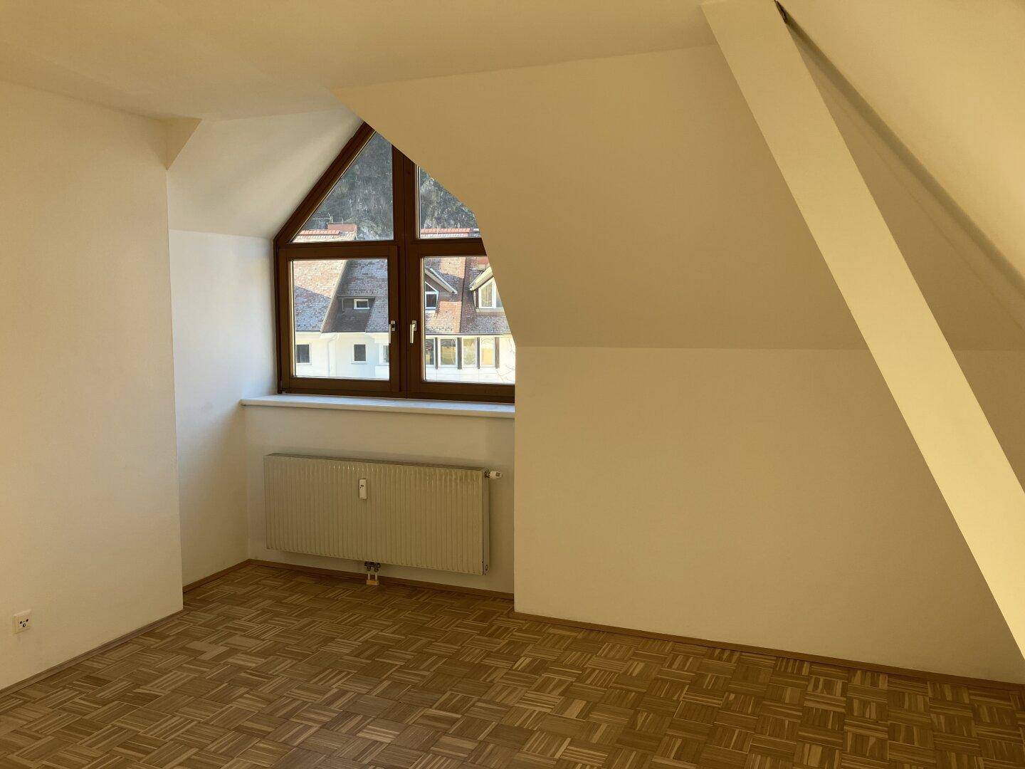 Unmöblierter Raum im Dachgeschoss mit Parkettboden, schrägen Wänden und Gaubenfenster.