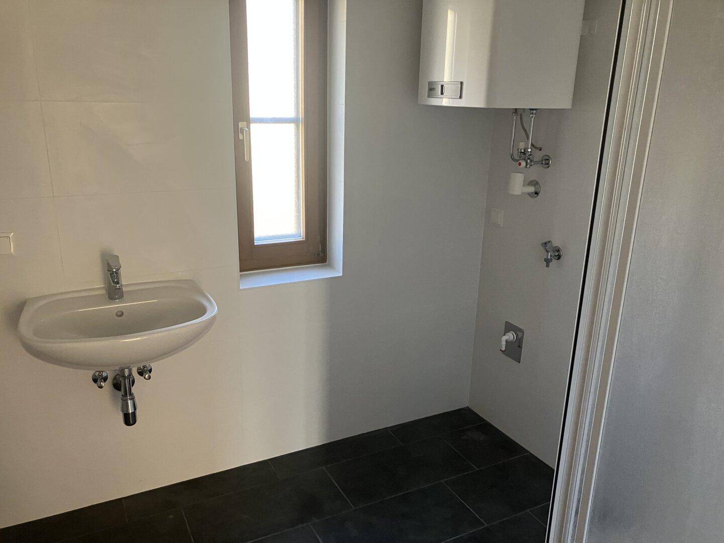 Helles Badezimmer mit Waschbecken, Fenster und Warmwasserboiler an der Wand.