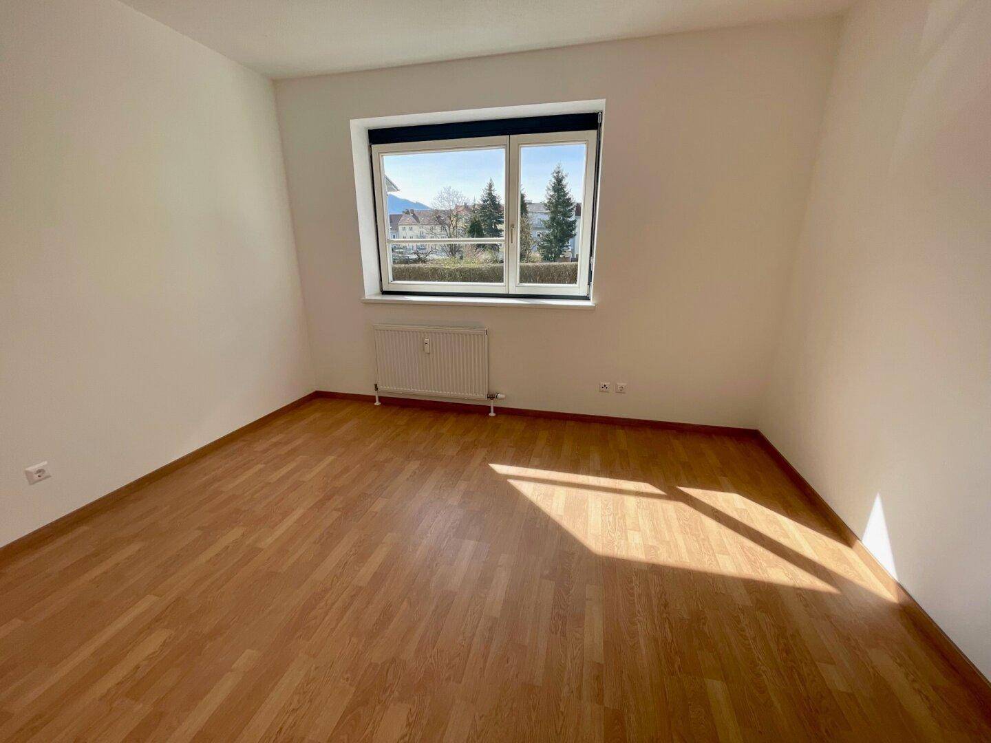 Freundliches Zimmer mit Laminatboden und Blick ins Grüne durch das Fenster.