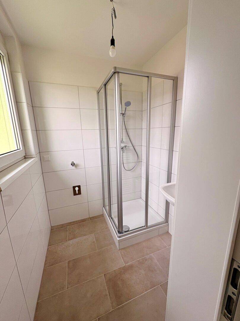 Helles Badezimmer mit Duschkabine, Fenster und hellen Fliesen an Wänden und Boden.