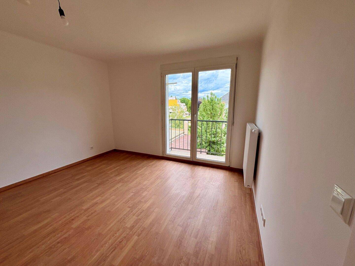 Heller Wohnraum mit Laminatboden, großem Fenster und Zugang zu einem französischen Balkon.