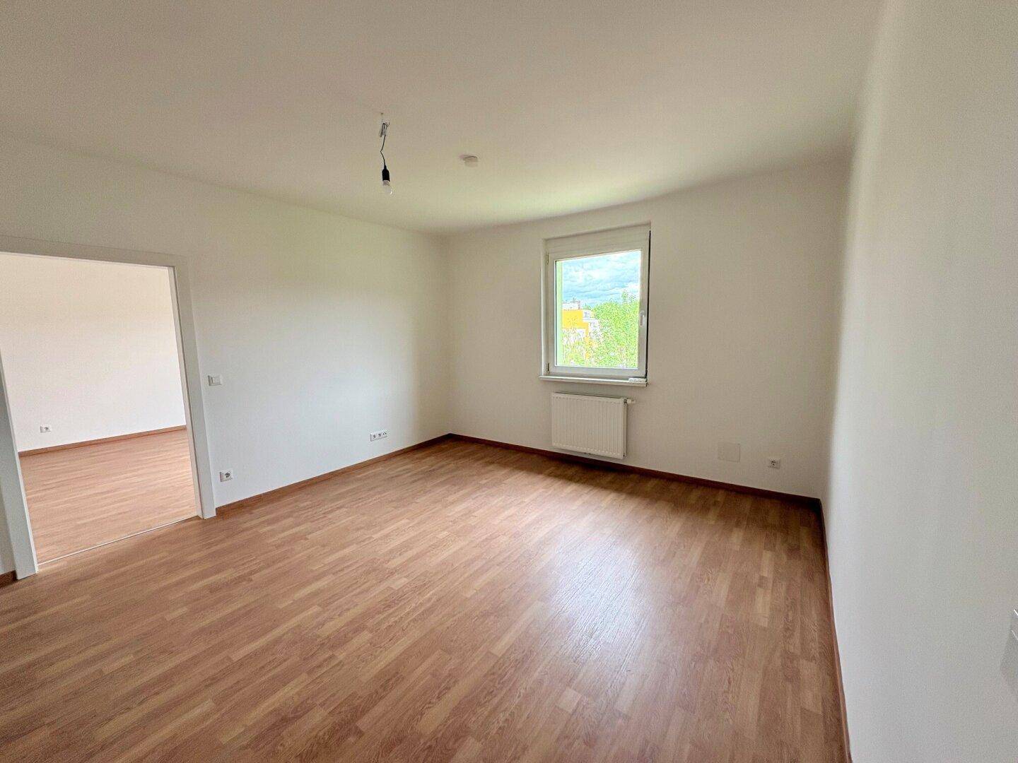 Geräumiger, heller Raum mit Laminatboden, Fenster und offenem Zugang zu einem weiteren Zimmer.