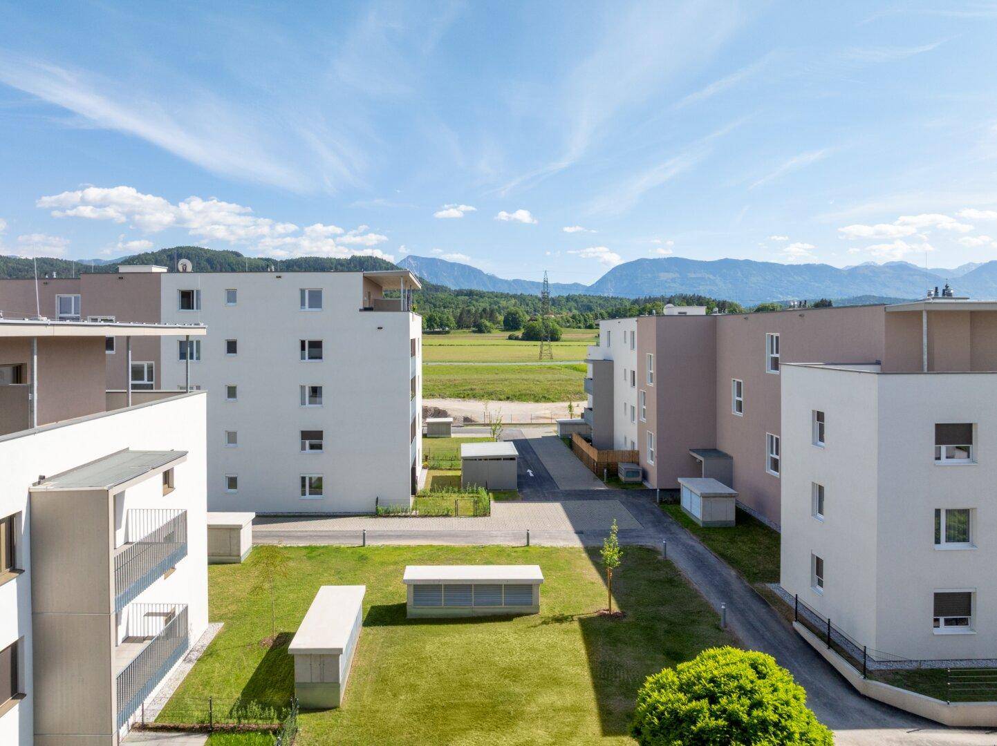 Wohnanlage mit modernen Gebäuden, umgeben von Grünflächen und Bergen in der Ferne unter klarem Himmel.