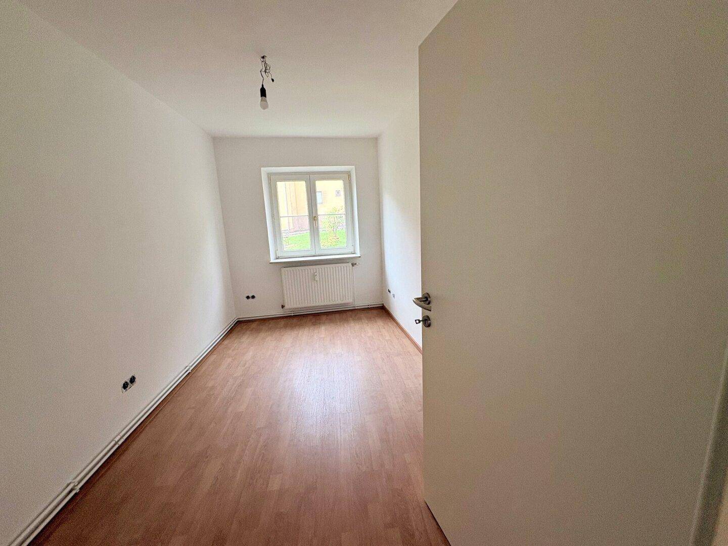 Helles Zimmer mit braunem Laminatboden und weiß gestrichenen Wänden, mit Blick ins Grüne.