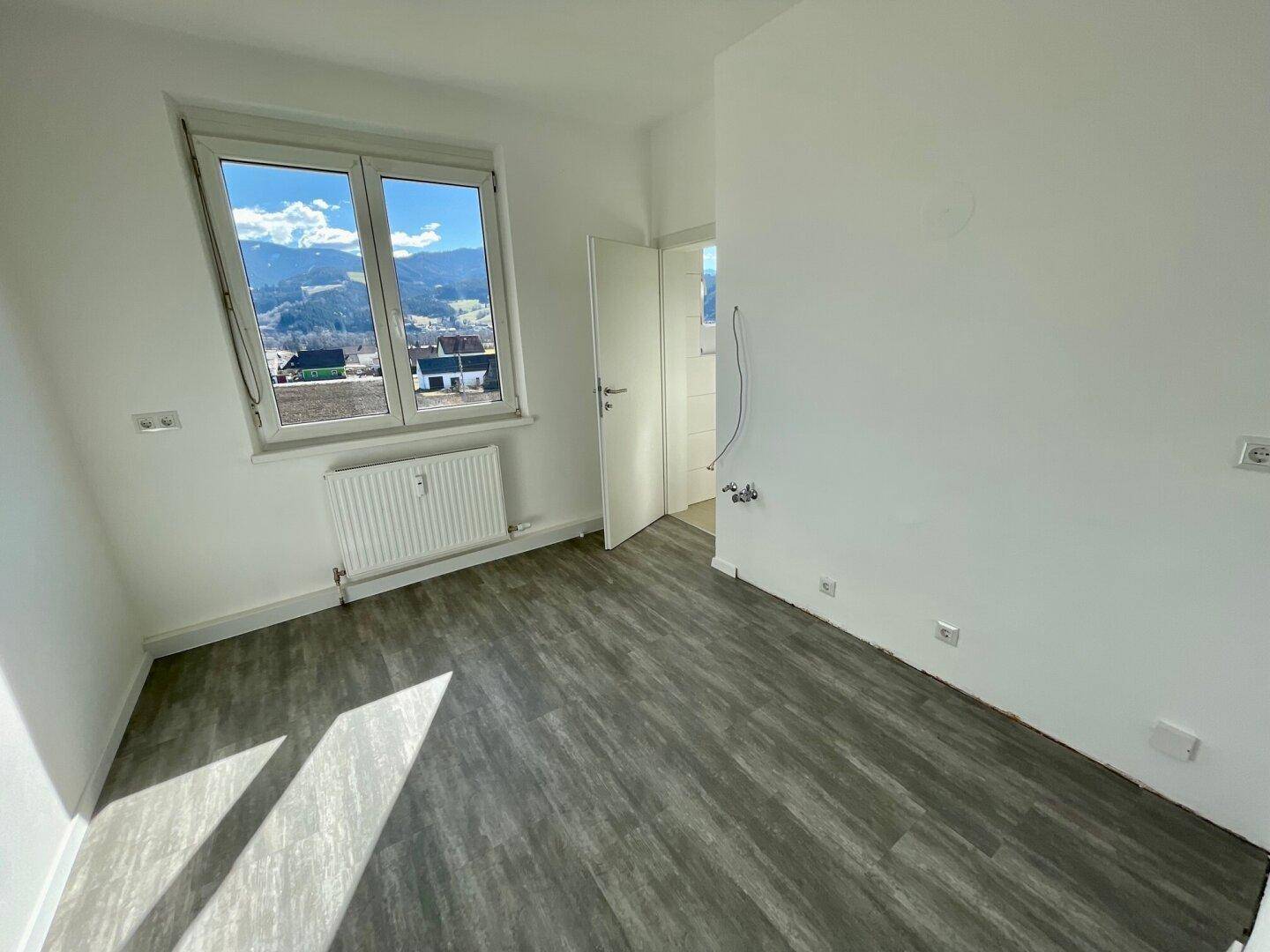 Kleineres Zimmer mit grauem Boden, Heizkörper und Fenster mit Bergblick, angrenzend an ein Badezimmer.