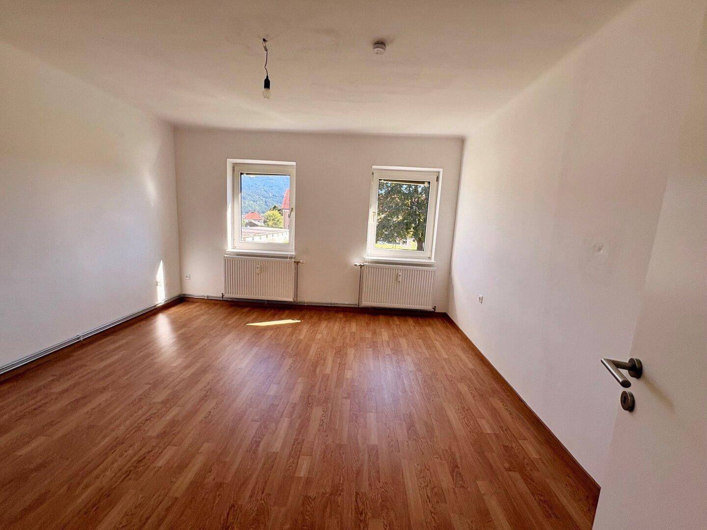 Heller, leerer Wohnraum mit Laminatboden und zwei Fenstern mit Bergblick.
