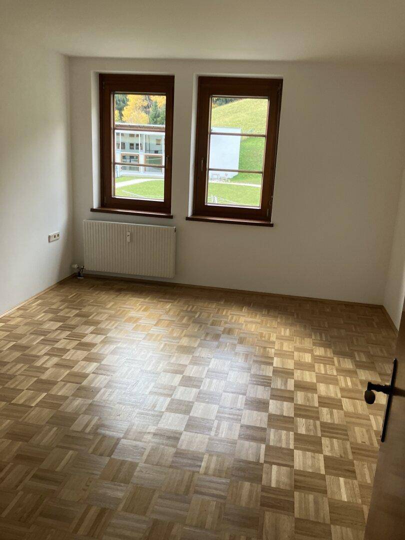 Leeres Zimmer mit Fischgrätparkettboden und zwei Fenstern mit Blick ins Grüne.