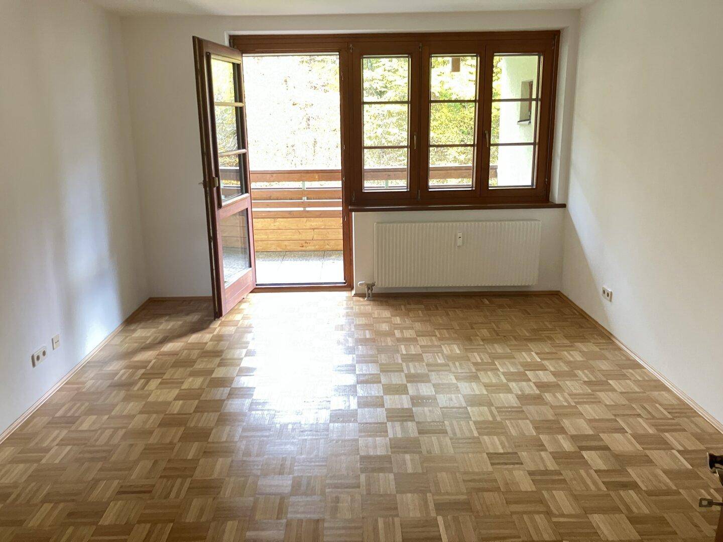 Helles Zimmer mit Parkettboden und Zugang zu einem Balkon, umgeben von grüner Natur.