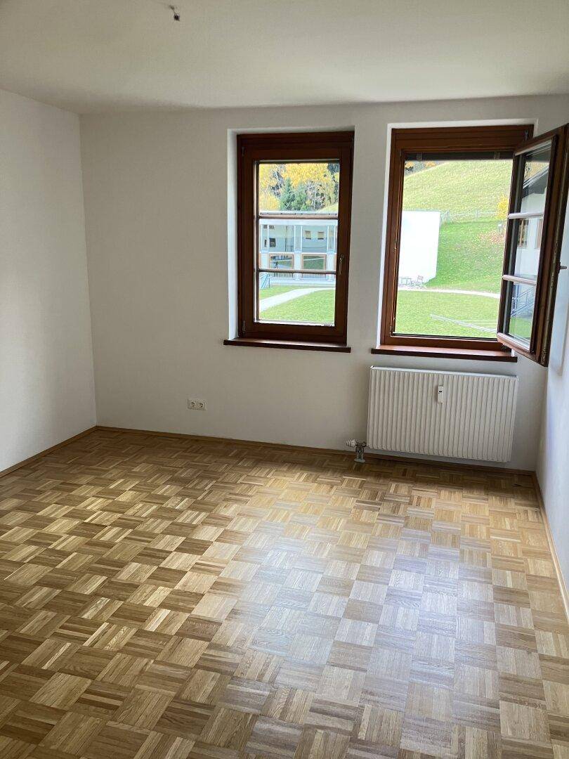 Helles Zimmer mit Parkettboden und zwei Fenstern, eines davon geöffnet, mit Blick ins Grüne.